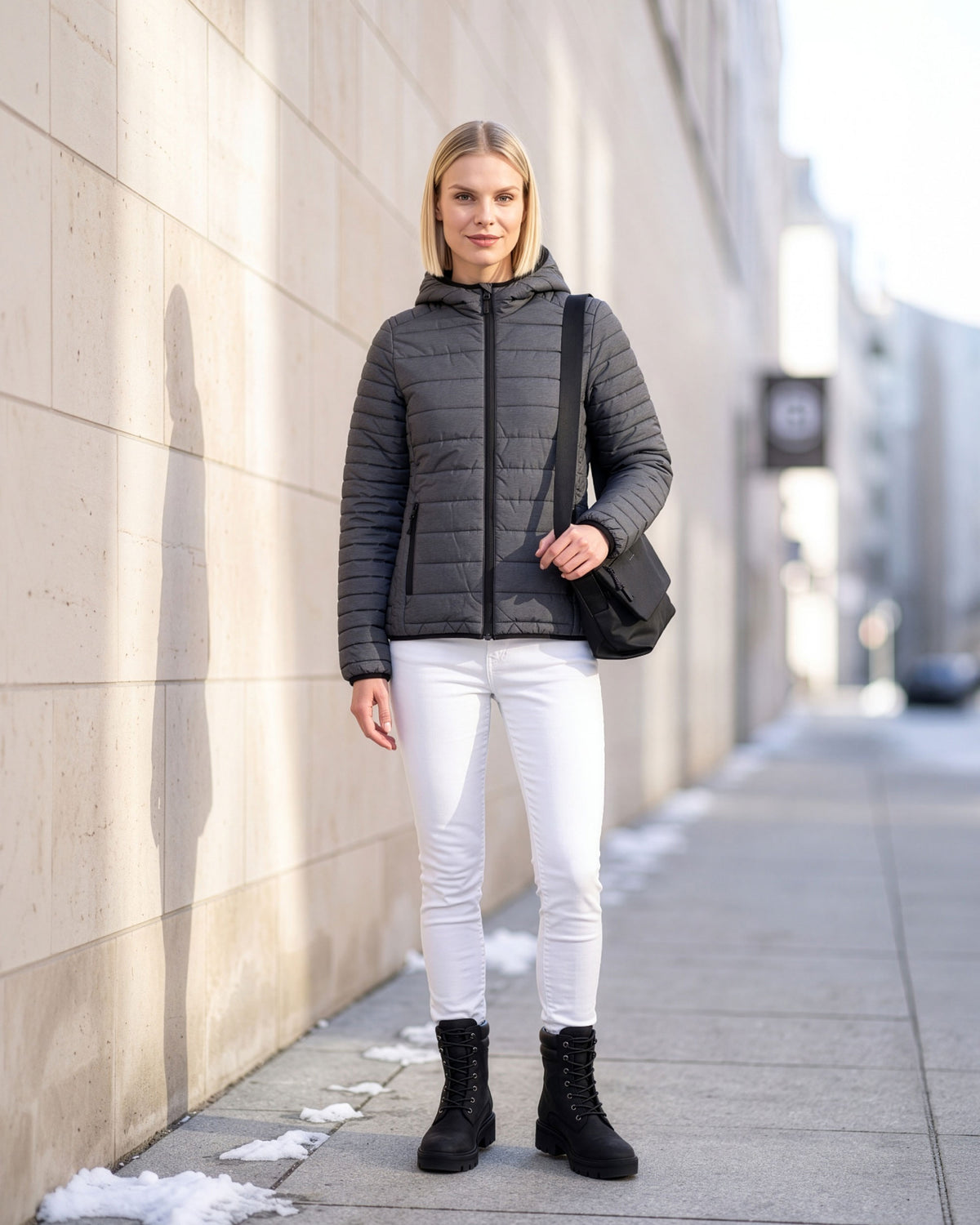 VIVID Schultertasche Damen schwarz mit grauer Kapuzen-Steppjacke, weißer Hose und schwarzen Schnürboots – Frau an einer hellen Steinwand auf einem Gehweg mit Schneeresten stehend