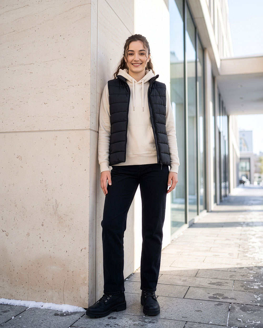 VIVID Hoodie Damen cremeweiß mit dunkelblauer Steppweste, schwarzer Hose und schwarzen Boots – Frau lächelnd an einer hellen Steinwand neben großen Glasfenstern und Schneeresten lehnend