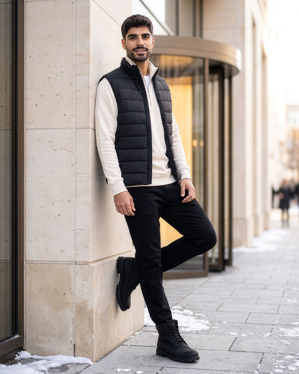 VIVID Sweatshirt Herren cremeweiß mit dunkelblauer Steppweste, schwarzer Hose und schwarzen Schnürboots – Mann mit einem Fuß an einer hellen Steinwand neben einer Drehtür und Schneeresten lehnend