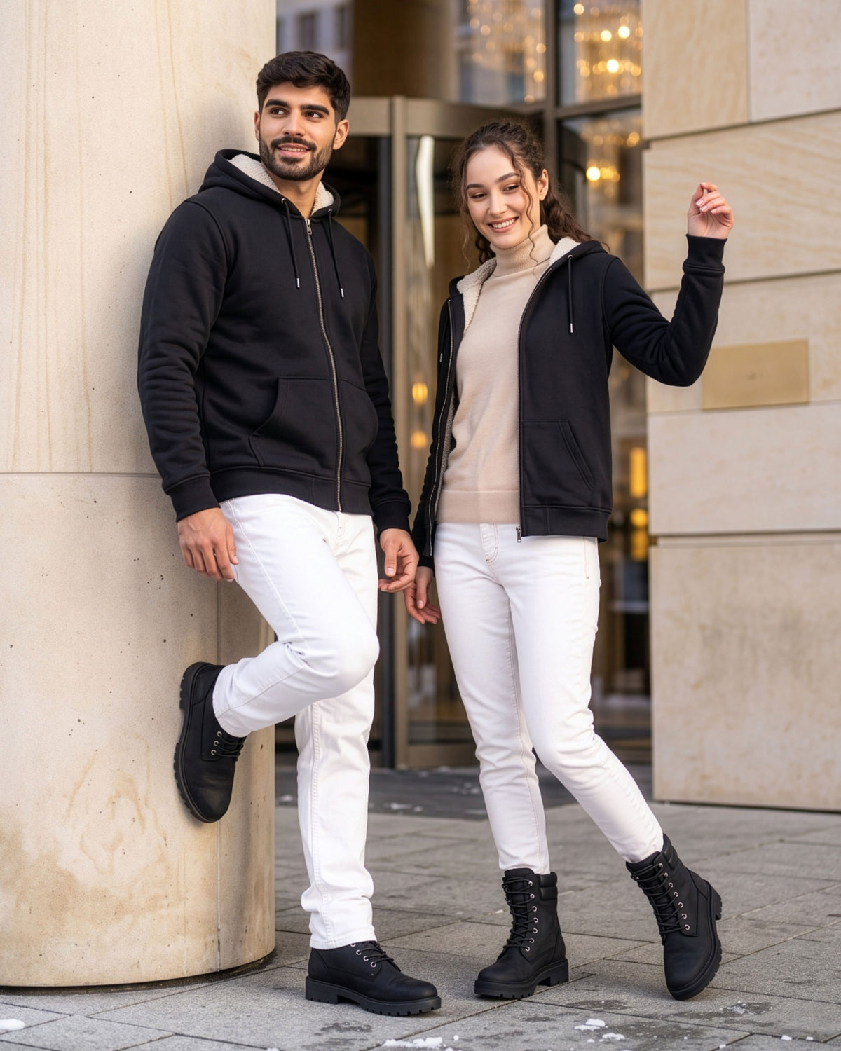 VIVID Unisex Zip-Hoodies schwarz mit hell gefütterten Kapuzen, weißen Hosen und schwarzen Boots – Paar lächelnd neben einer hellen Steinsäule und Drehtür mit Schneeresten, Frau mit beigem Rollkragenpullover