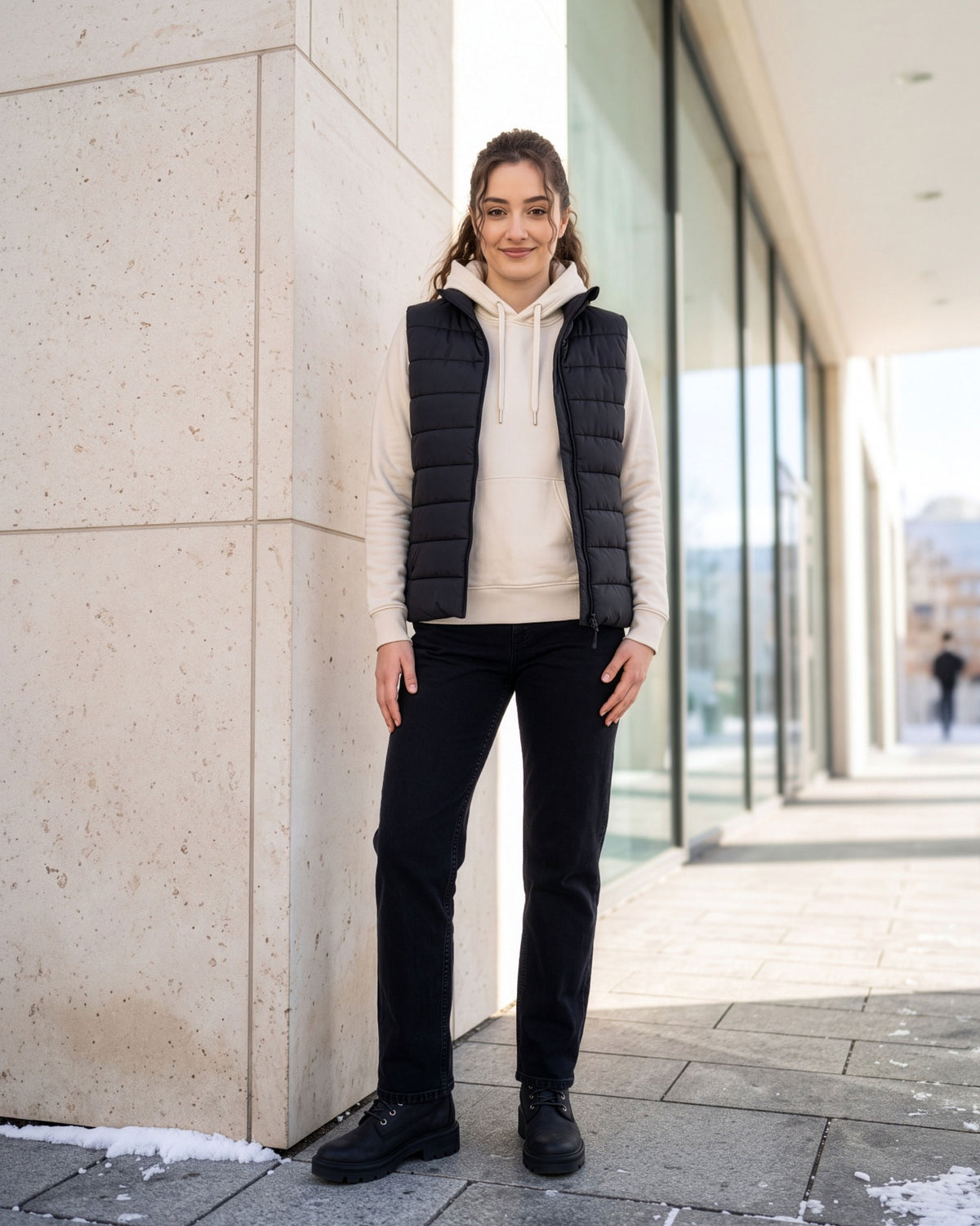VIVID Hoodie Damen cremeweiß mit dunkelblauer Steppweste, schwarzer Hose und schwarzen Boots – Frau lächelnd an einer hellen Steinsäule neben großen Glasfenstern und Schneeresten stehend