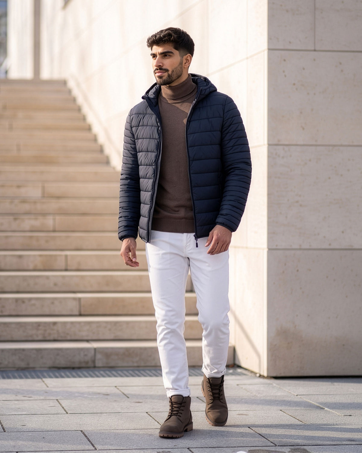 VIVID Hose Herren weiß mit dunkelblauer Kapuzen-Steppjacke, braunem Rollkragenpullover und braunen Schnürboots – Mann vor einer hellen Steintreppe gehend und zur Seite blickend