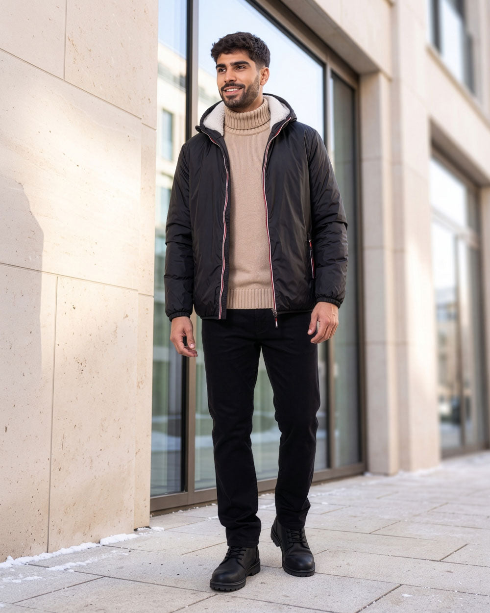 VIVID Winterjacke Herren schwarz mit hell gefütterter Kapuze, beigem Rollkragenpullover, schwarzer Hose und schwarzen Boots – Mann lächelnd vor einer hellen Gebäudefassade mit großen Fenstern und Schneeresten gehend