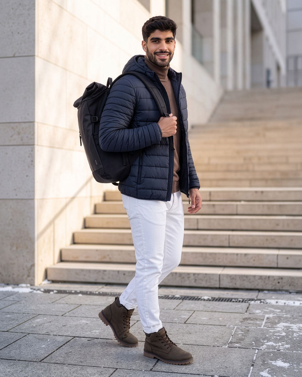 VIVID Rolltop-Rucksack schwarz – Mann mit dunkelblauer Kapuzen-Steppjacke, braunem Rollkragenpullover, weißer Hose und braunen Schnürboots trägt den Rucksack vor einer hellen Steintreppe mit Schneeresten
